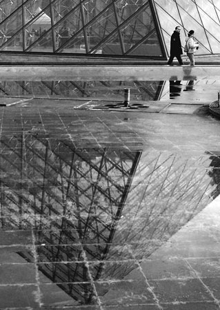 dans un des bassins &agrave; moiti&eacute; vide, le reflet fragment&eacute; de la pyramide du Louvre, un couple passe dans le fond. Noir et blanc