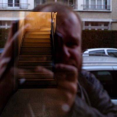Reflet de face du photographe sur le verre fumé d'une porte vitrée et, visible de l'autre côté de cette porte, d'un escalier vivement éclairé par un rayon de soleil. Il semble nous mener droit dans la tête du photographe.